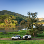 Two white suvs parked on a dirt road near the water.